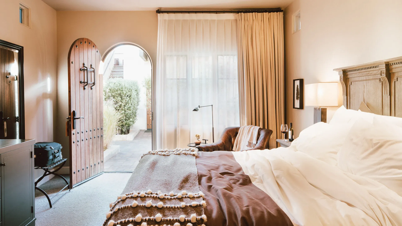 A beautiful bedroom features a large bed with white sheets and a brown comforter in front of a curtained window and an open archway door | Hotel Yountville