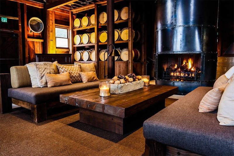 A cozy wine tasting room featuring comfortable seating, a rustic wooden table, and a warm fireplace, with wine barrels stacked against the wall creating an inviting atmosphere.