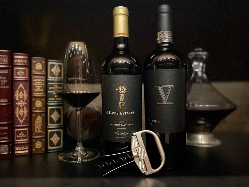 Two bottles of Davis Estates wine, one 2016 Cabernet Sauvignon and one 2018 Phase V, accompanied by a wine glass and corkscrew, set against a backdrop of classic books.