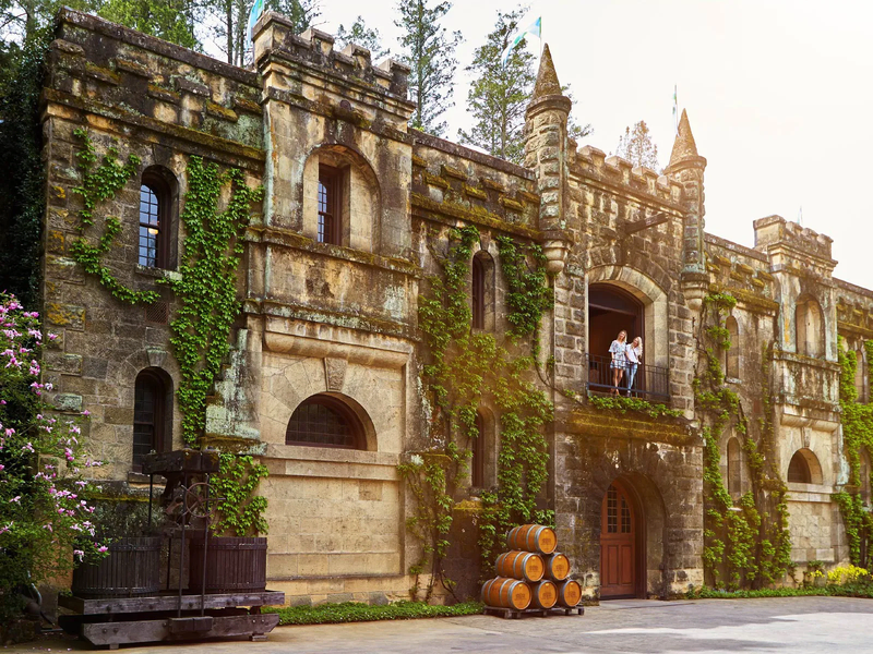 Historic stone winery building with ivy, barrels, and visitors on a balcony.