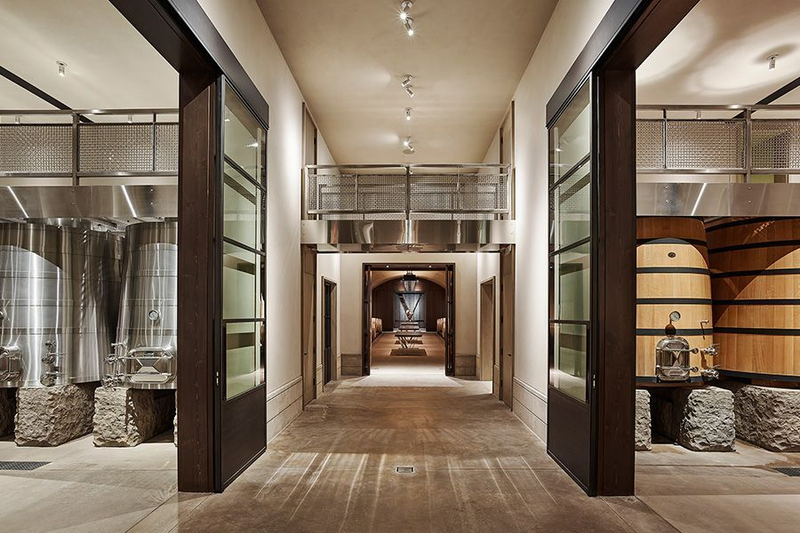 A modern winery interior featuring a wide hallway with stainless steel fermentation tanks on the left and large wooden barrels on the right. The design is sleek and industrial.