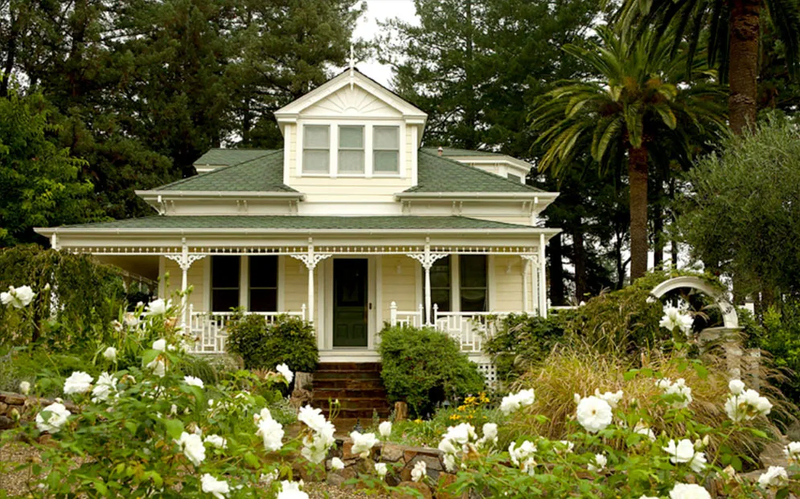 A charming, vintage-style house with a white exterior and green roof, surrounded by lush greenery and blooming white flowers.