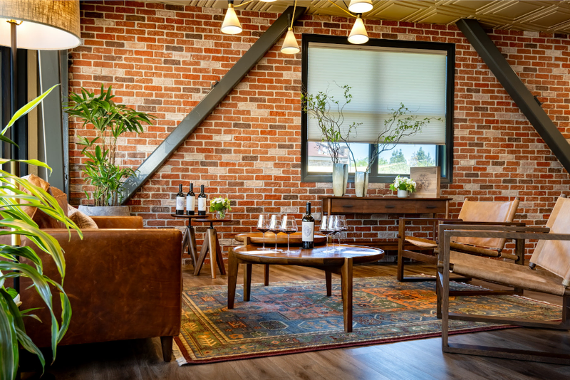 A cozy lounge with exposed brick walls, warm leather furniture, and a wooden coffee table. Wine bottles and glasses are arranged, adding a relaxed, elegant ambiance.