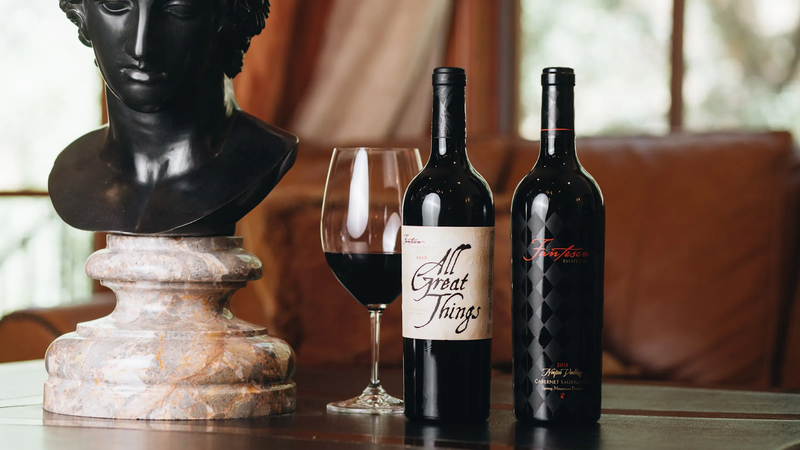 Two elegant wine bottles, one labeled "All Great Things," and the other "Fortune," are placed beside a glass of red wine and a marble bust, creating a refined setting.