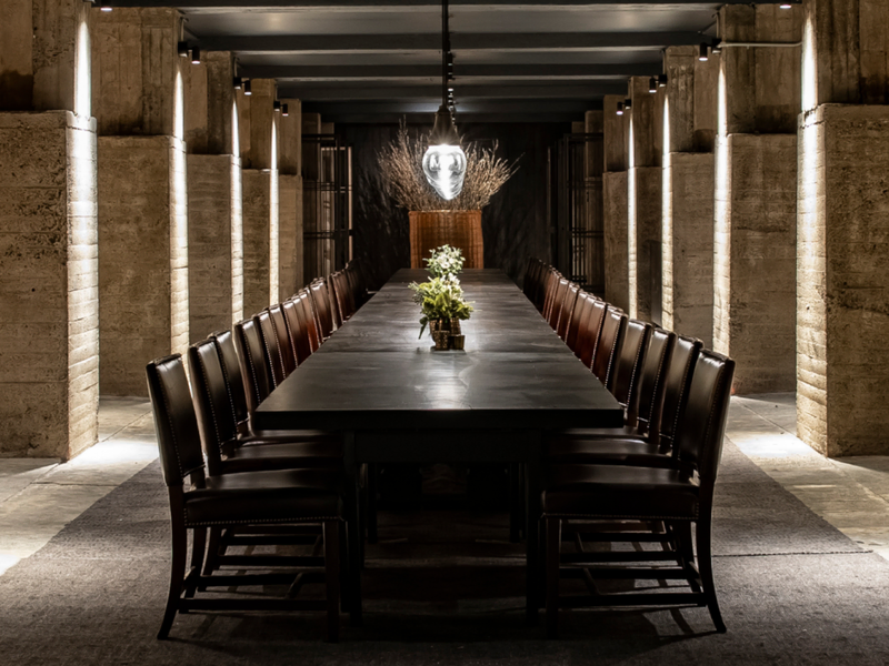  A long, elegant dining table with leather chairs is set in a dimly lit room with stone columns. A central light fixture and greenery create a refined, intimate atmosphere.