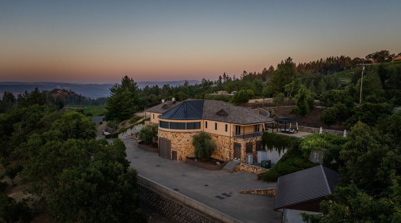  A stunning hillside property surrounded by lush greenery, featuring a stone building with a modern circular design. The sunset casts a warm glow over the serene landscape.