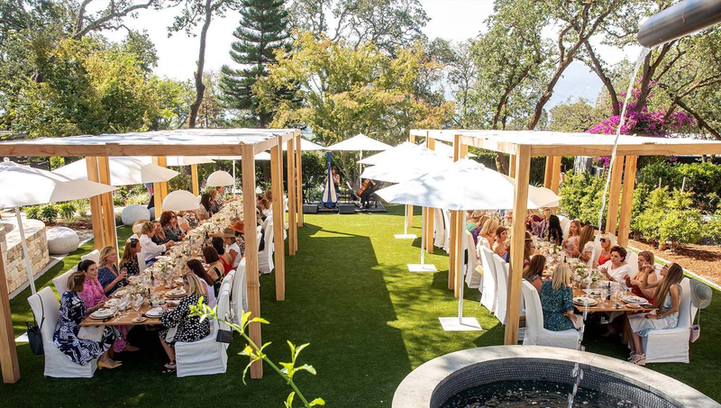 Elegant outdoor brunch with guests under wooden pergolas and white umbrellas.